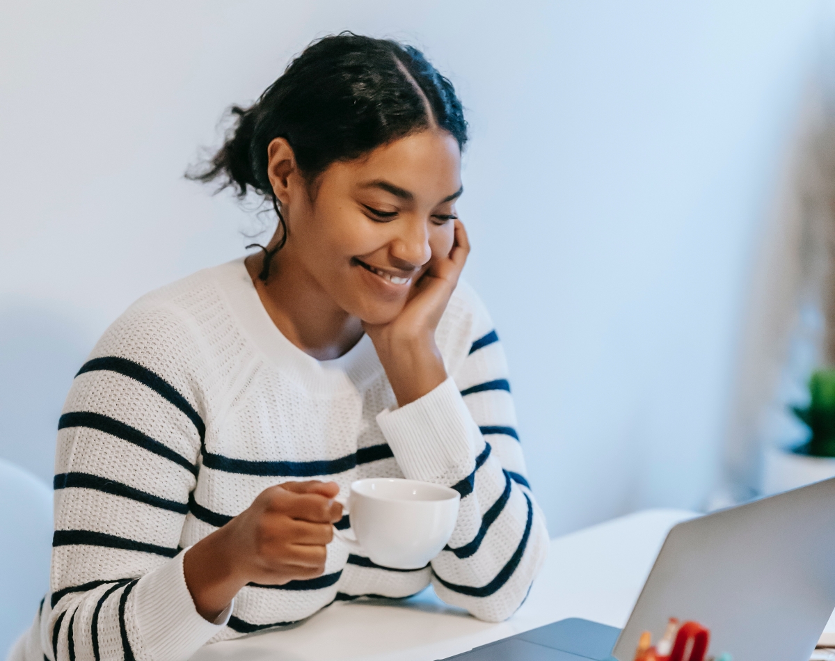 A smiling woman in a striped sweater holding a coffee cup while working on her laptop, representing a virtual assistant in a relaxed and productive workspace.