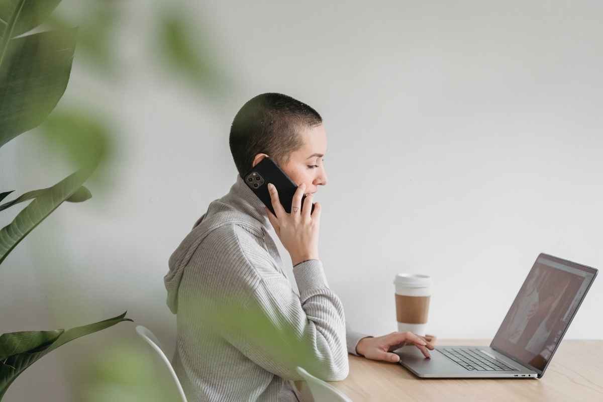 A woman in a gray hoodie is engaged in virtual communication, talking on the phone while working on her laptop at a desk with a takeaway coffee cup.