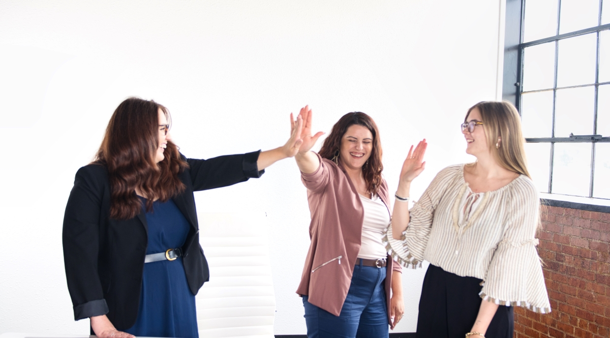 Three professional women smiling and high-fiving in a bright office space.