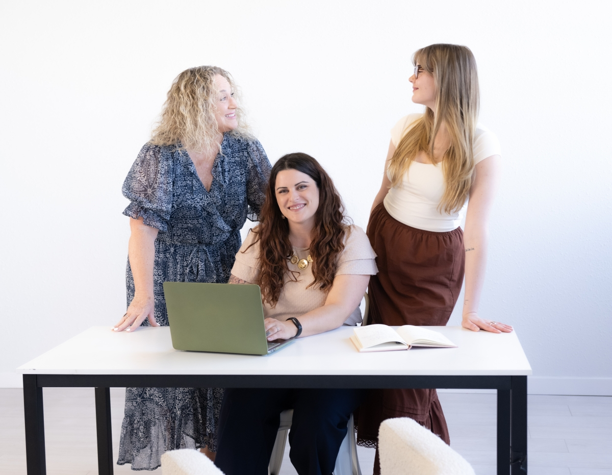 Three women collaborating at a table, with a laptop and notebook, representing teamwork and finding the best virtual assistant solutions.