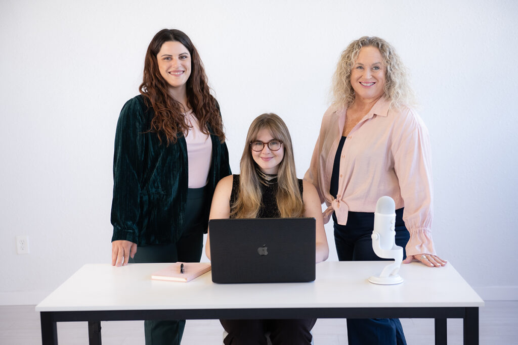 A team of three professionals from a virtual assistant agency, with one seated at a desk and two standing, all smiling confidently.