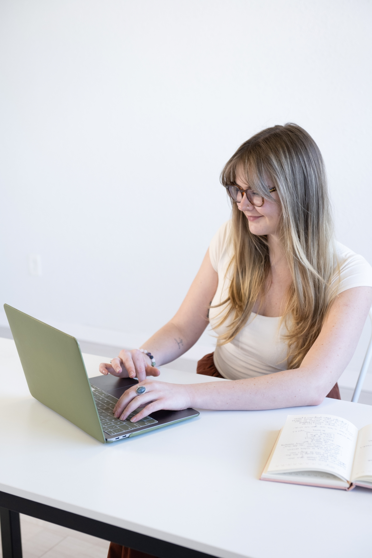 A woman with glasses, working on a green laptop at a desk, with an open notebook beside her.