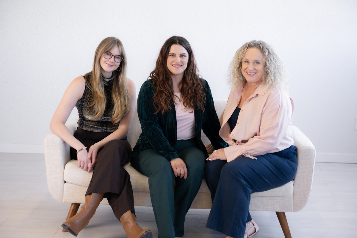 Three women sitting on a beige couch, smiling and looking at the camera. They are dressed in a mix of casual and semi-formal attire, with expressions of camaraderie and support.