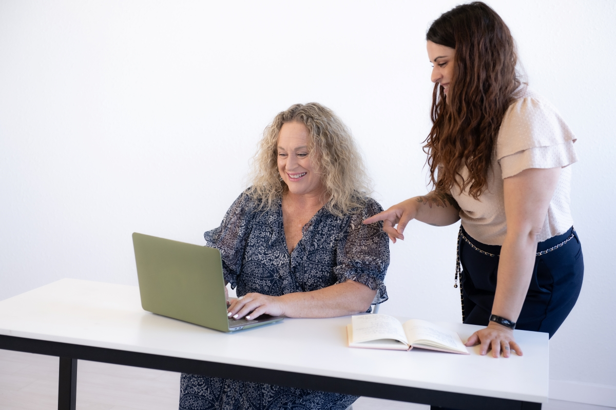 Two women working together, one sitting at a desk with a laptop while the other stands beside her pointing at the screen, emphasizing processes and workflows.