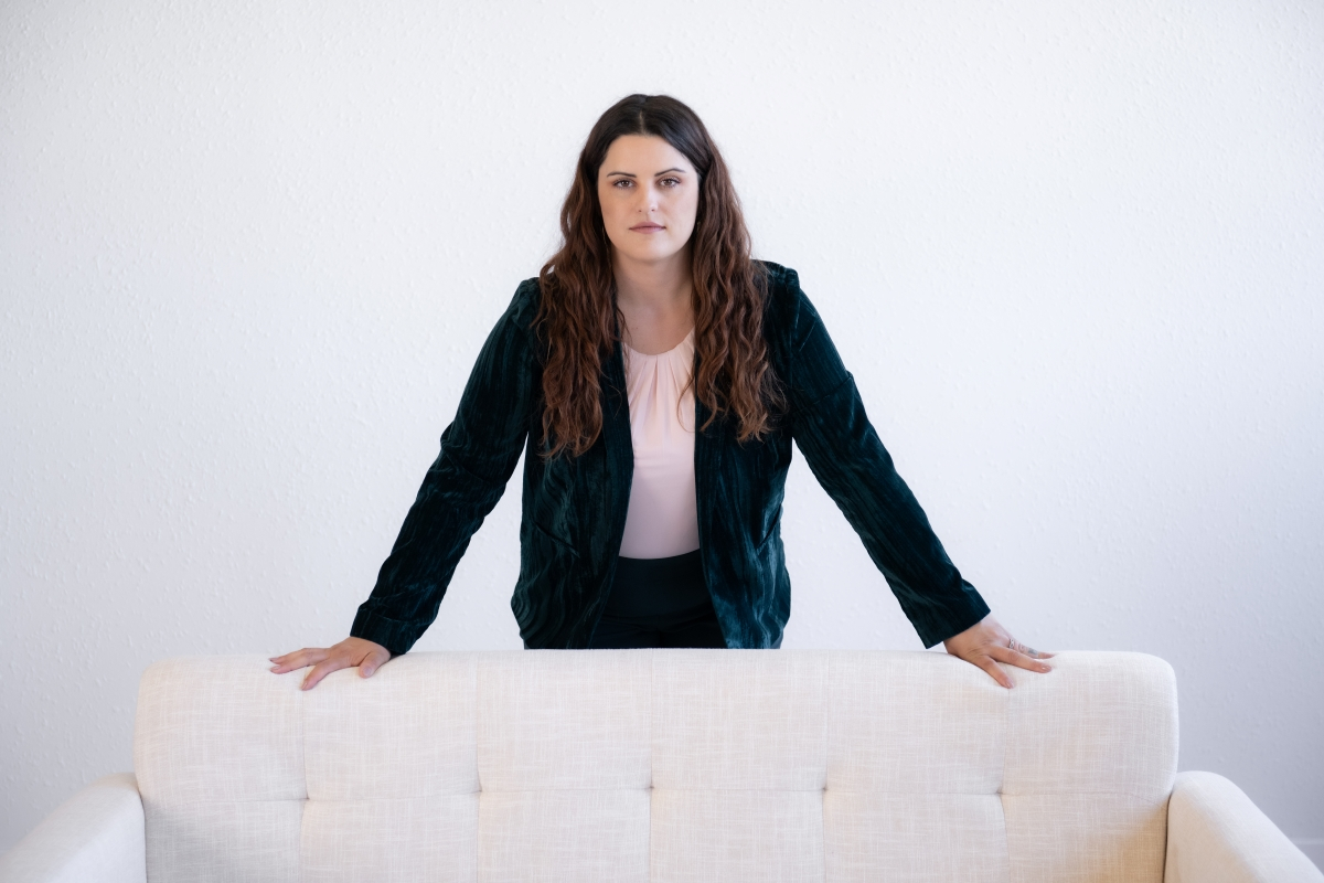 Confident woman standing behind a white couch, dressed in a dark green blazer and light pink blouse, against a minimalist white background.