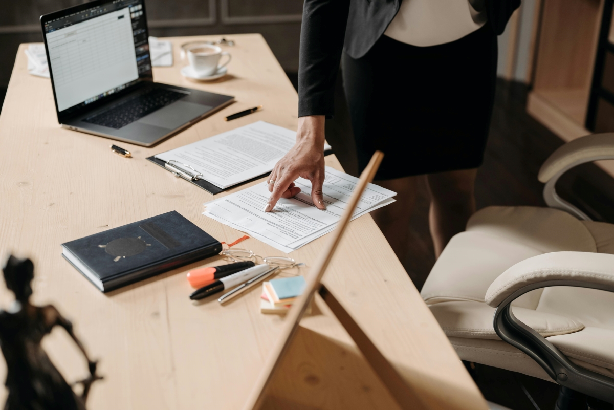 Close-up of a professional pointing to a document on a desk, surrounded by contracts, policies, a laptop, and office supplies.