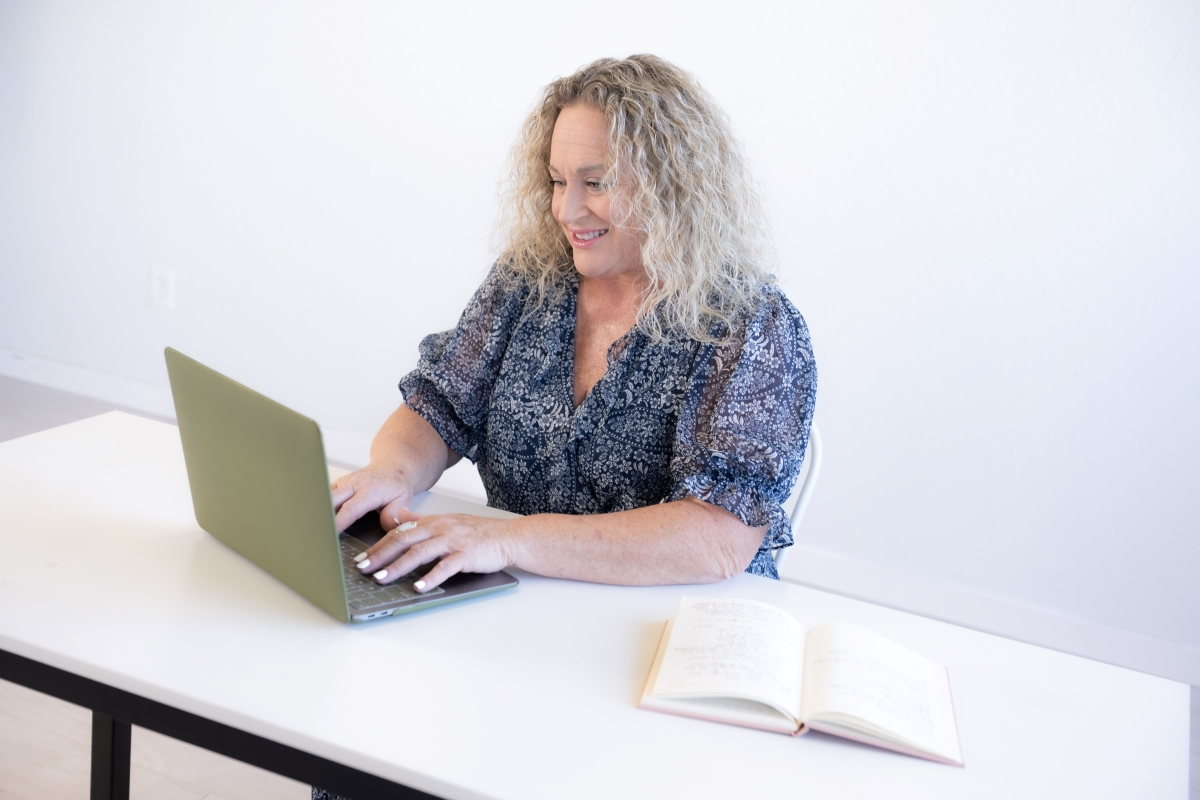 A professional woman with curly blonde hair working on a laptop at a white desk, with an open notebook beside her.