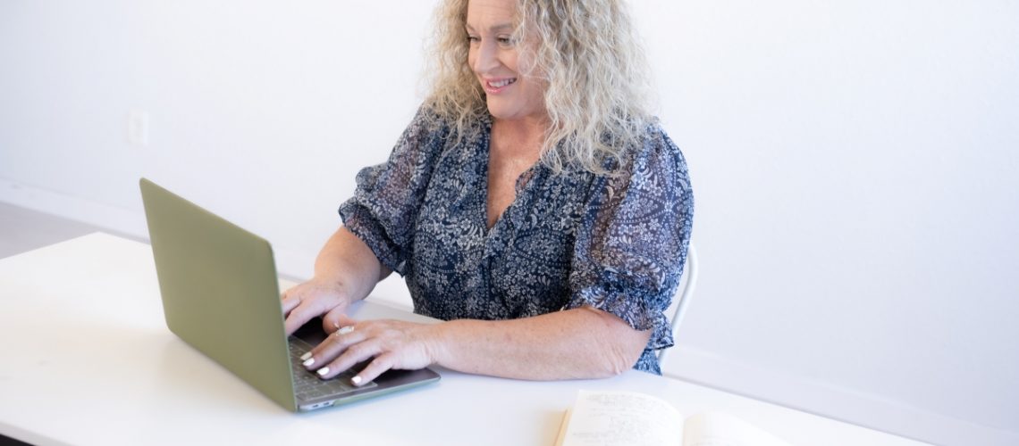 Smiling woman with curly hair typing on a green laptop at a desk with an open journal, representing productivity and VA tips for success.