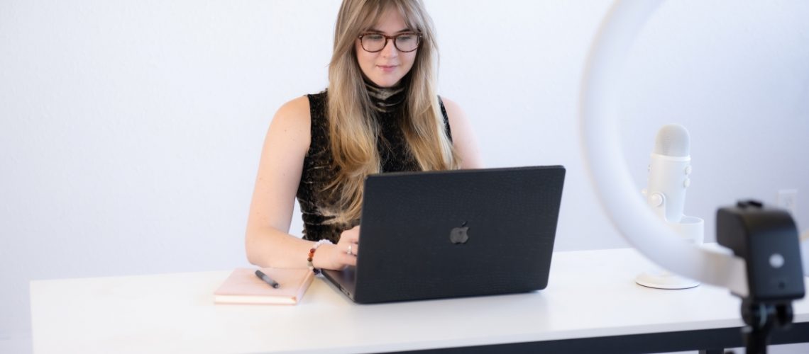 Focused woman with long blonde hair and glasses, working on a laptop with a microphone and ring light, setting herself up for success this fall.