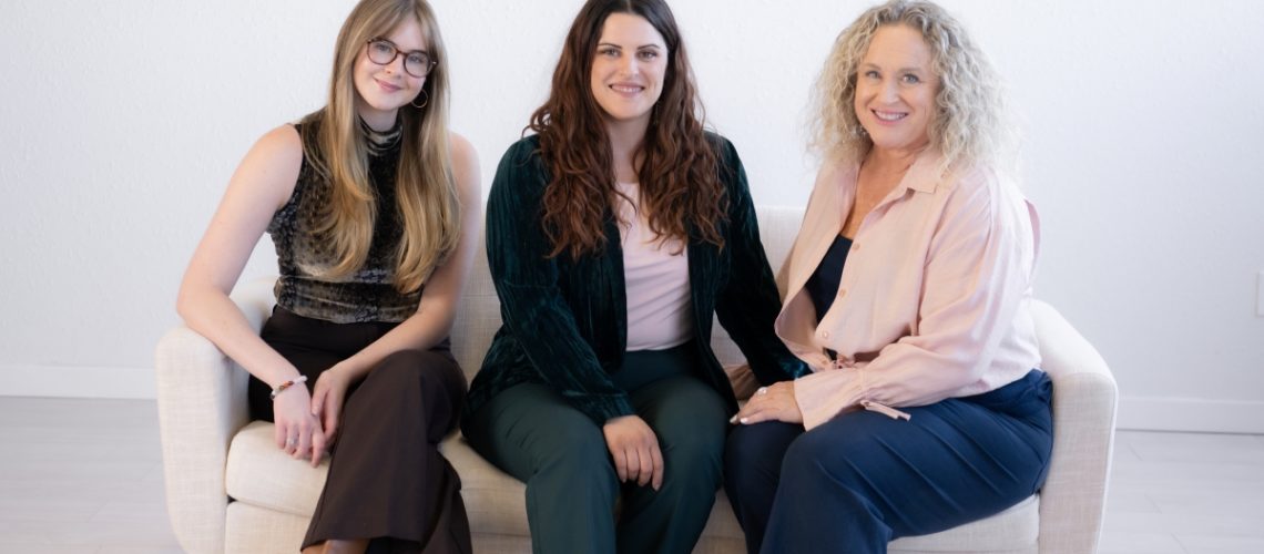 Three women sitting on a beige couch, smiling and looking at the camera. They are dressed in a mix of casual and semi-formal attire, with expressions of camaraderie and support.