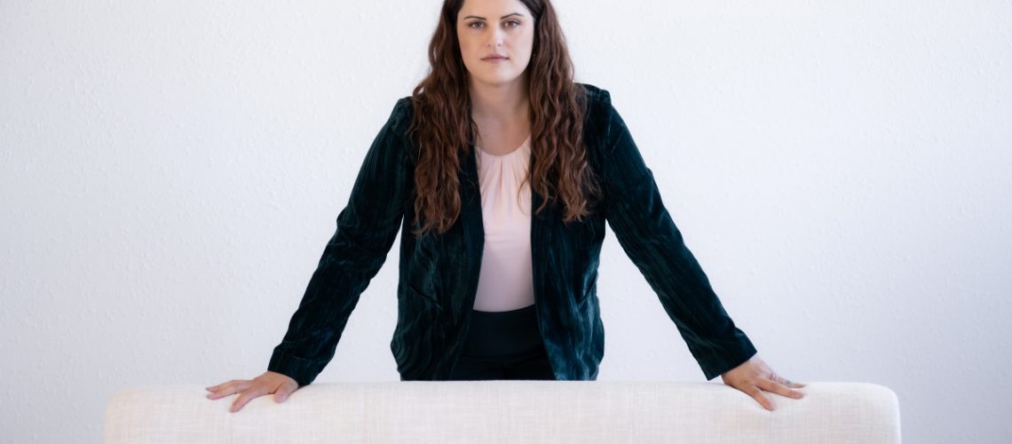 Confident woman standing behind a white couch, dressed in a dark green blazer and light pink blouse, against a minimalist white background.