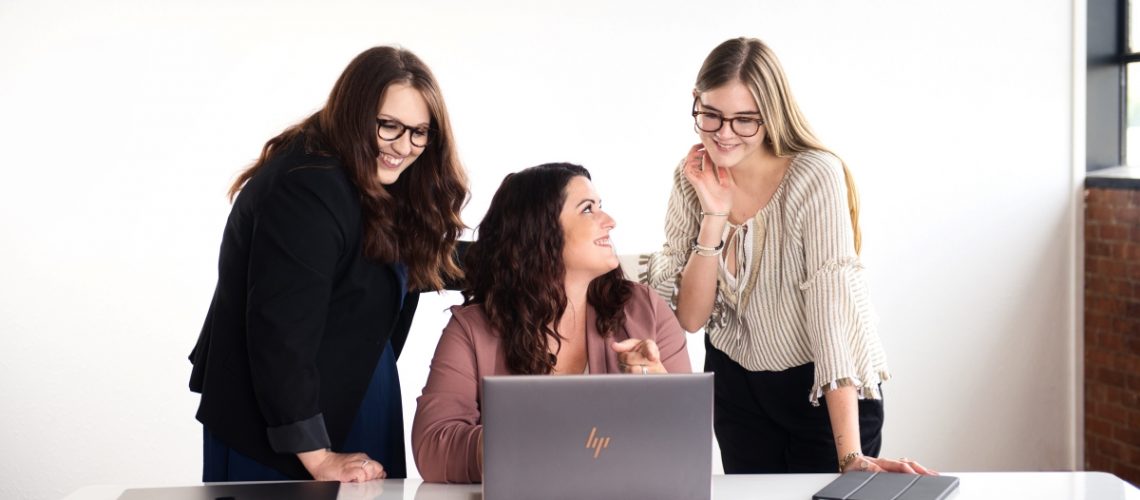 A group of three women collaborating at a desk, focused on a laptop, working together to achieve business goals.