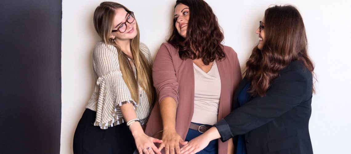 Three smiling women standing together with their hands stacked, symbolizing teamwork and collaboration while planning for the holidays.