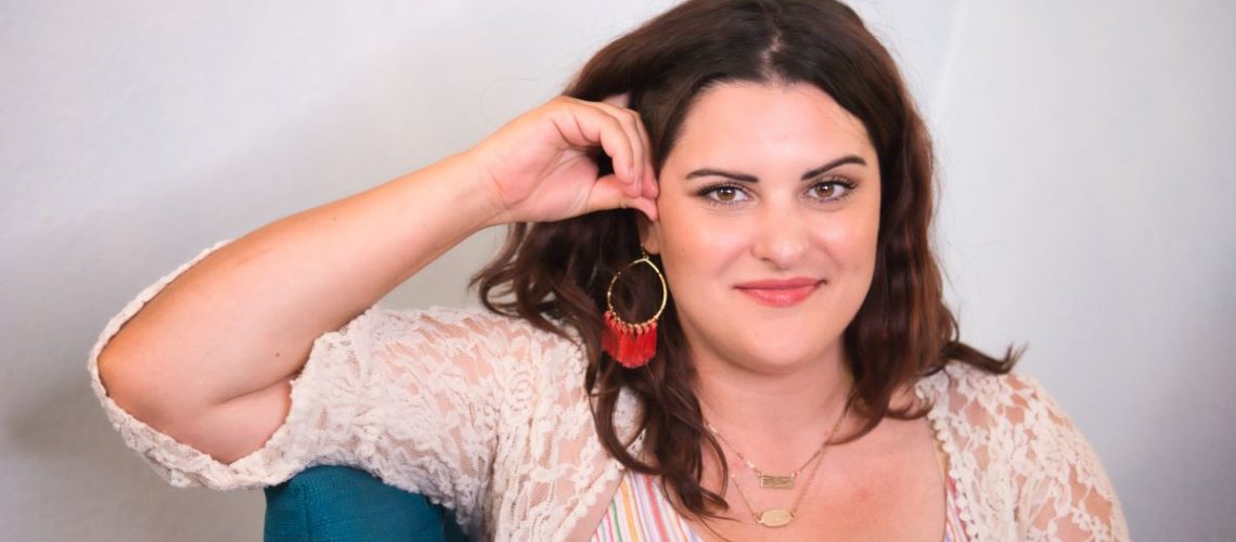 A confident woman with wavy brown hair, wearing a lace cardigan and colorful earrings, smiling at the camera.