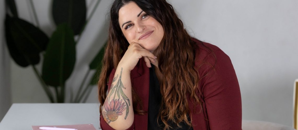 Smiling woman with long wavy hair, wearing a maroon blazer, sitting at a desk with notebooks—representing a professional virtual assistant ready to help scale your business.