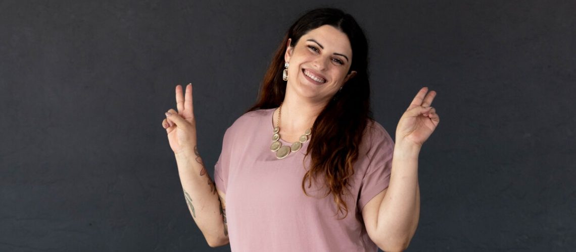 A smiling woman in a pink blouse and white pants poses with peace signs in front of a dark gray wall, featured on Canvas Rebel.