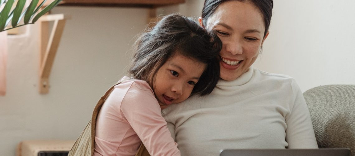 A smiling mother and her young daughter sitting together on a couch, enjoying time on a laptop, representing warmth and togetherness during the holiday season.