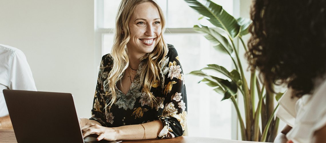 A smiling woman with a laptop in a business meeting, engaging in a discussion with colleagues.