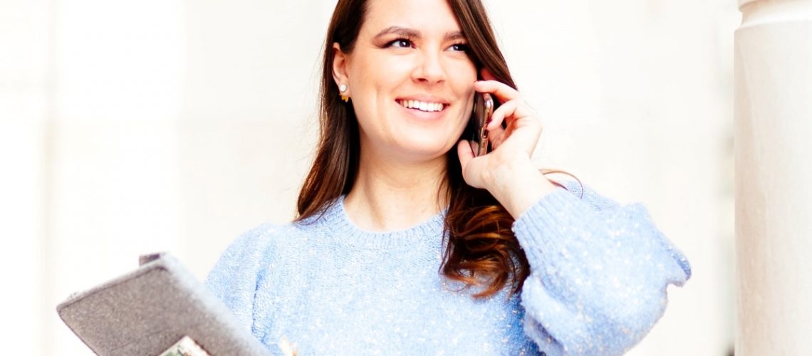 A smiling woman in a blue sweater talking on the phone while holding a notebook and pen, representing confidence and working at your own pace.