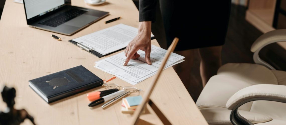 Close-up of a professional pointing to a document on a desk, surrounded by contracts, policies, a laptop, and office supplies.