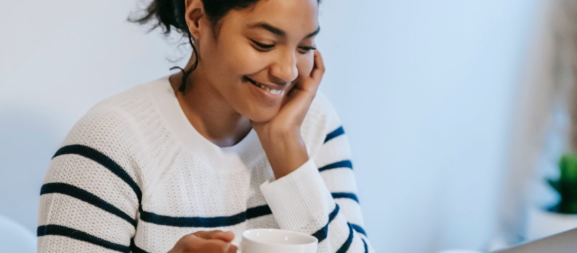 A smiling woman in a striped sweater holding a coffee cup while working on her laptop, representing a virtual assistant in a relaxed and productive workspace.